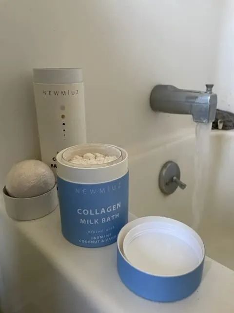 A bottle of collagen milk bath and a bath ball sit near a running faucet, suggesting a serene, relaxing bath experience.