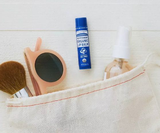 A canvas pouch containing a makeup brush, pink sunglasses, a blue tube of peppermint organic lip balm, and a spray bottle, all displayed on a light-colored surface.