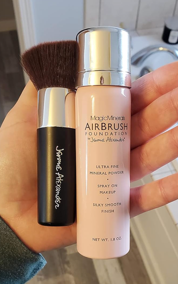 A hand holding both a brush and a bottle of MagicMinerals AirBrush Foundation by Jerome Alexander, promising an ultra-fine, smooth finish.
