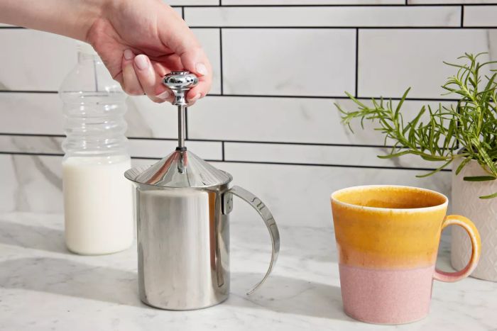 A hand gripping the knob of the HIC Fino Milk Frother, positioned beside a carafe of milk and a mug
