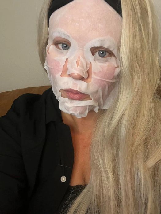 A person with long hair wearing a sheet mask, gazing at the camera, dressed casually in a black top