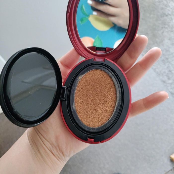 A hand holds an open compact foundation with a cushion makeup sponge, while another hand, adorned with painted nails, holds what seems to be a phone or notebook in the background.