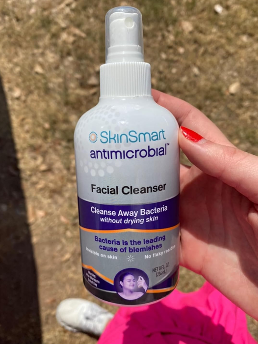 A person holding a bottle of SkinSmart Antimicrobial Facial Cleanser, with text highlighting its effectiveness at cleansing bacteria while maintaining moisture in the skin.