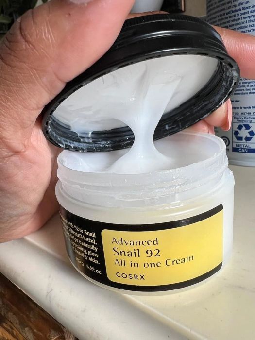 A person holds an open jar of COSRX Advanced Snail 92 All-in-One Cream, with the cream’s texture clearly visible.