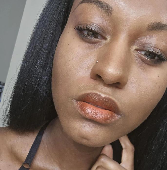 Close-up of a person with smooth skin, wearing minimal makeup and a black top, gently holding their chin while posing for a beauty or fashion feature