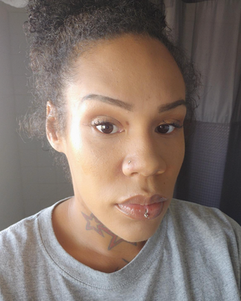 A person with a star neck tattoo and lip piercing, wearing a gray shirt, staring directly at the camera.
