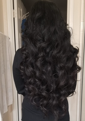 A person with long, wavy black hair stands in a doorway, showing off the length and style of their hair.