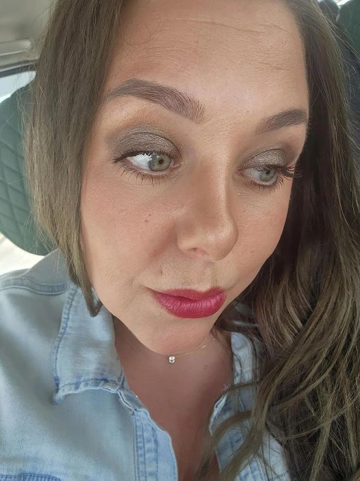 A person wearing a denim shirt, sporting subtle eye makeup and a bold lip color, takes a close-up selfie.
