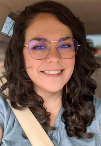 A person with glasses and a seatbelt, smiling in a selfie with long, wavy hair, dressed in casual attire.