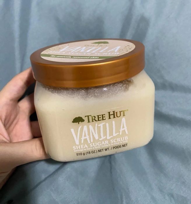 A hand holding a jar of Tree Hut Vanilla Shea Sugar Scrub on a soft gray fabric.