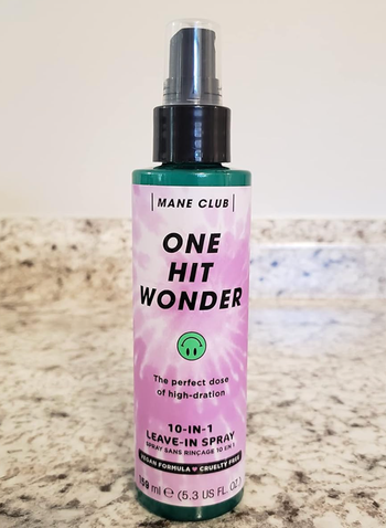 A bottle of Mane Club One Hit Wonder 10-in-1 Leave-In Spray sitting on a marble countertop. The product is known for its high hydration properties and is both vegan and cruelty-free.