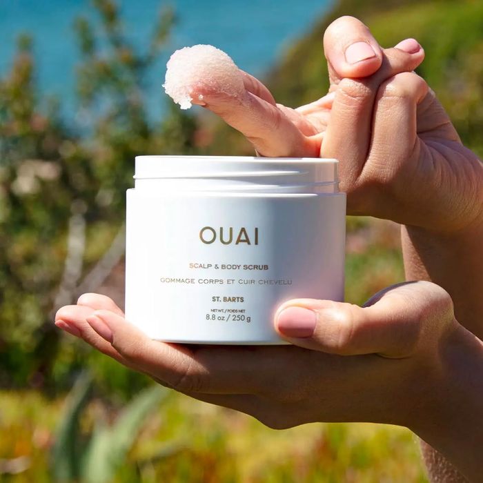 A person holding a jar of the OUAI Scalp and Body Scrub with a dab of the product on their finger against a natural backdrop
