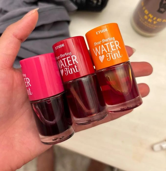 Three mini bottles of Etude Dear Darling Water Tint in pink, red, and orange, held in a hand and placed on a light table