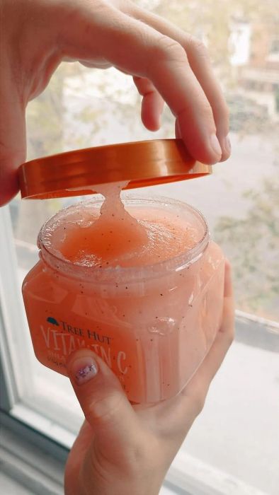 Hands opening a jar of Tree Hut Vitamin C sugar scrub, positioned near a bright window.