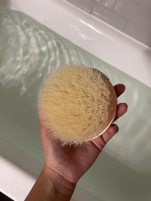A hand holding a round bath brush with natural bristles, positioned next to a filled bathtub, ready for use.