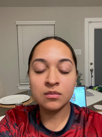 A person sits indoors with closed eyes, wearing a casual top, with a table containing a laptop and some plates in the background.
