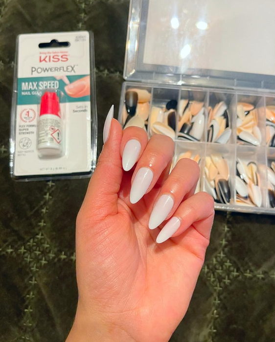 A hand showcasing long, white, coffin-shaped nails with a package of KISS PowerFlex nail glue and a box of assorted fake nails in the background.