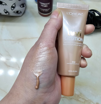 A hand holding the L'Oréal liquid foundation tube, with the product dripping onto the skin, showing its smooth texture and consistency for easy shopping decisions.