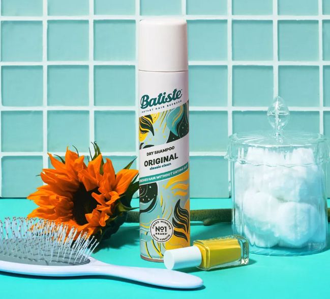 Batiste Original Dry Shampoo placed on a tiled surface next to a hairbrush, a sunflower, a bottle of nail polish, and a jar filled with cotton balls.