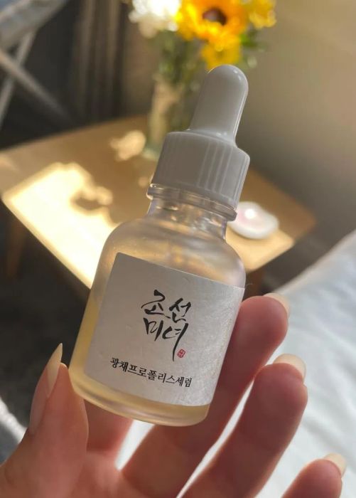 A dropper bottle of beauty serum with Korean text on the label, held in a hand indoors with flowers softly blurred in the background