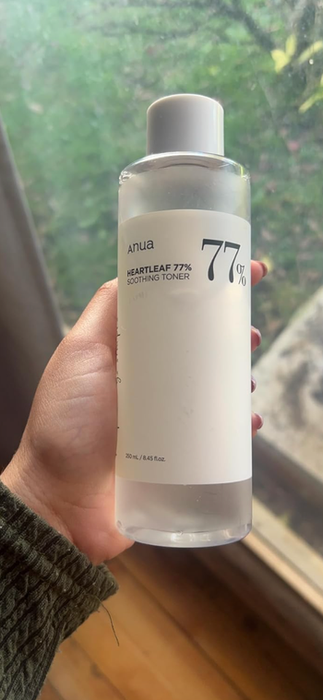 A hand holding a bottle of Anua Heartleaf 77% Soothing Toner, with a background featuring an indoor setting, part of a window, and greenery outside.