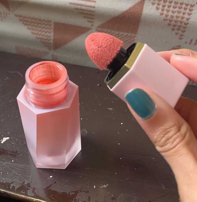 A person unscrewing the cap of a liquid blush container, which has a built-in sponge applicator, set against a decorative background.