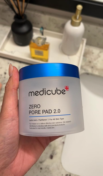 A hand holding the Medicube Zero Pore Pad 2.0 container, a skincare product designed to treat pores, set against a bathroom background with other bathroom items.