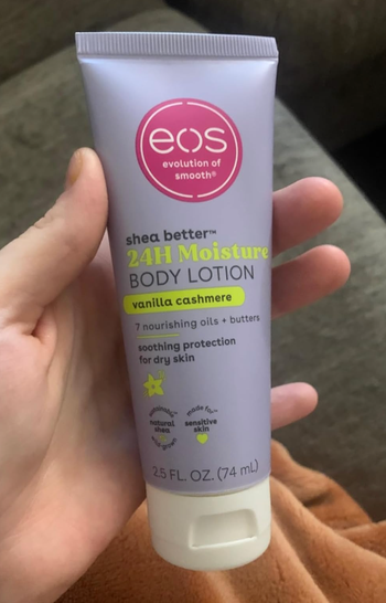 A hand holds a tube of EOS 24H Moisture Body Lotion in Vanilla Cashmere, with a focus on its soothing and protective qualities for dry skin