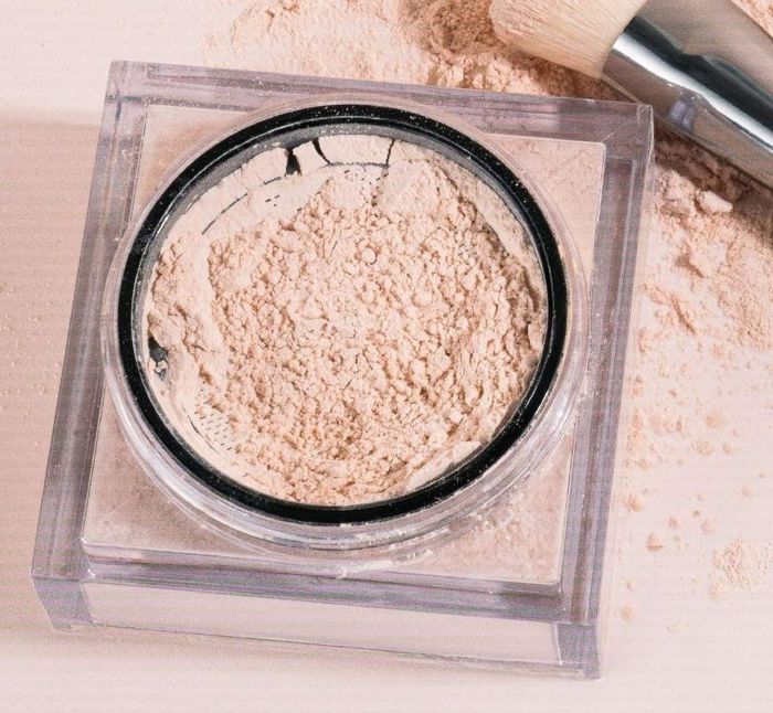 A container of loose face powder with a makeup brush resting beside it on a wooden surface
