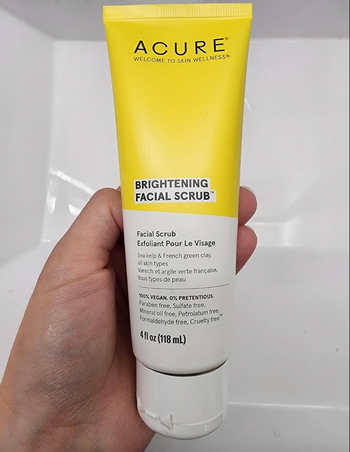A hand holding a tube of ACURE Brightening Facial Scrub.