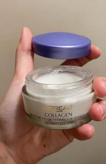 A hand holding an open jar of L'Oréal Collagen Moisture Filler Day-Night cream with its lid removed, revealing the product inside.