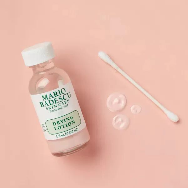 A bottle of Mario Badescu Drying Lotion with a cotton swab and droplets on a simple background