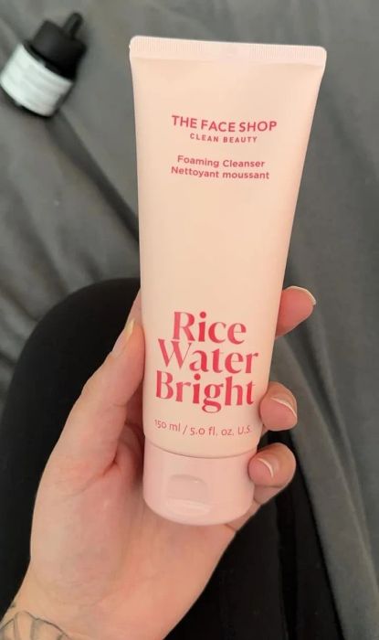 A hand holding The Face Shop's Rice Water Bright Foaming Cleanser tube, with a skincare dropper bottle visible in the background