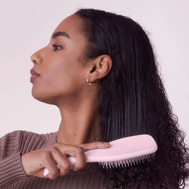 A model with long, dark, wavy hair uses a pink brush to comb through her wet locks.