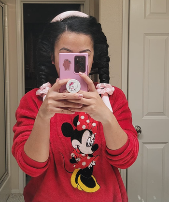 A person takes a mirror selfie, dressed in a red sweater featuring a Minnie Mouse print and holding a pink phone with a cute grip.