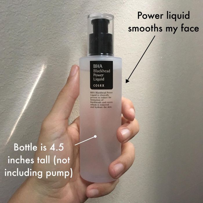 A hand holds a 4.5-inch tall bottle of COSRX BHA Blackhead Power Liquid, with text explaining its role in smoothing the face by clearing away blackheads.