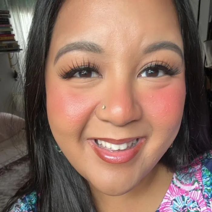 A person beams while wearing a floral-patterned top, with a sofa and partially drawn curtains visible in the background
