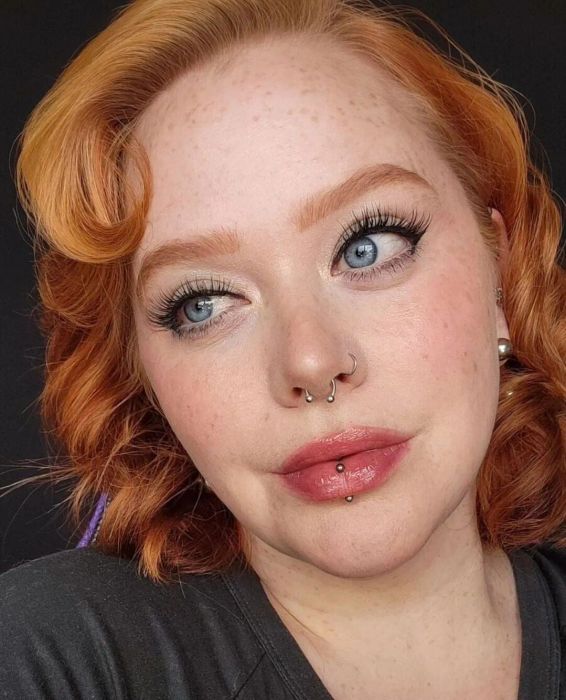 A person with red hair and piercings gazes thoughtfully, wearing minimal makeup and a simple top.