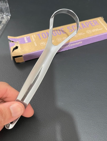 Stainless-steel tongue scraper displayed over a table with its packaging in the background.