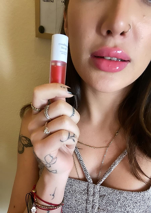 The reviewer holding the appleberry lip oil while showcasing its glossy effect on their lips.
