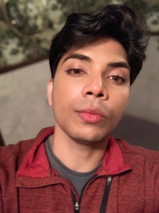 A person taking a selfie indoors, dressed casually in a red and gray jacket, with a neutral expression on their face