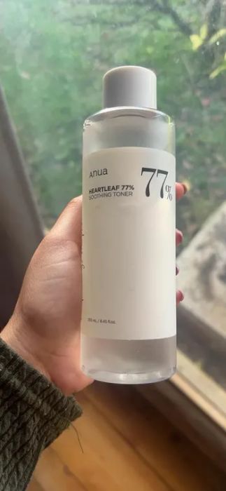A hand holding a bottle of Anua Heartleaf 77% Soothing Toner, with an indoor setting in the background featuring part of a window and lush greenery outside.