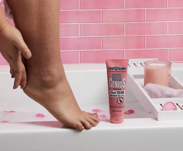 A person applying Soap & Glory Heel Genius foot cream to their foot while seated on a bathtub edge, with a tray holding a candle and towel beside them.