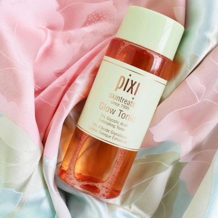 A bottle of Pixi Glow Tonic placed on a pastel-patterned fabric backdrop, highlighting its 5% glycolic acid content and exfoliating properties.