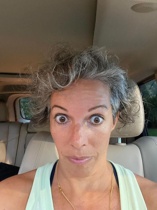A reviewer with curly, frizzy gray hair, wide eyes, and a look of surprise, sitting in a car while wearing a sleeveless top