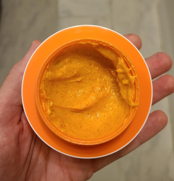 Reviewer holding an open jar of a vibrant orange face mask