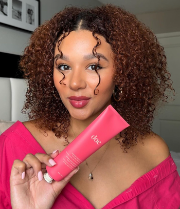 A model with curly hair holding a tube of hair product.