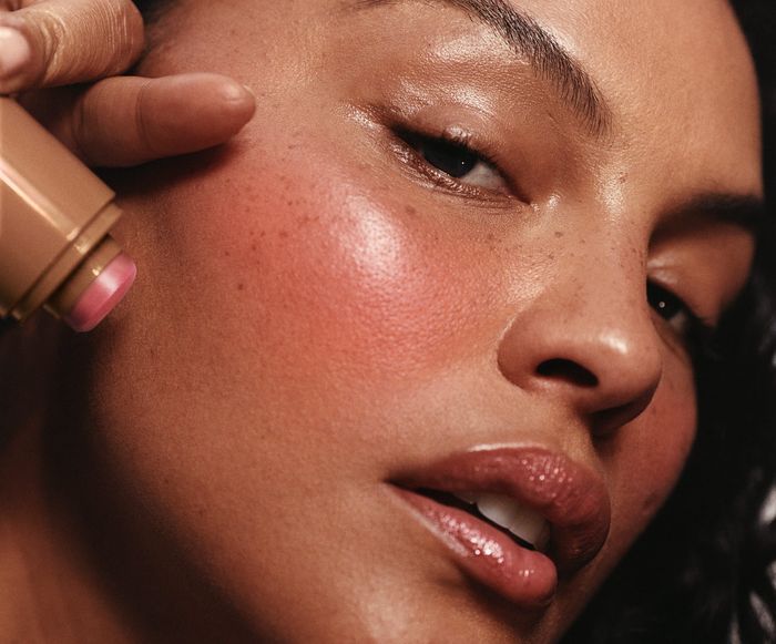 A close-up of someone applying a cream blush stick to their cheek, showing off smooth skin and natural freckles