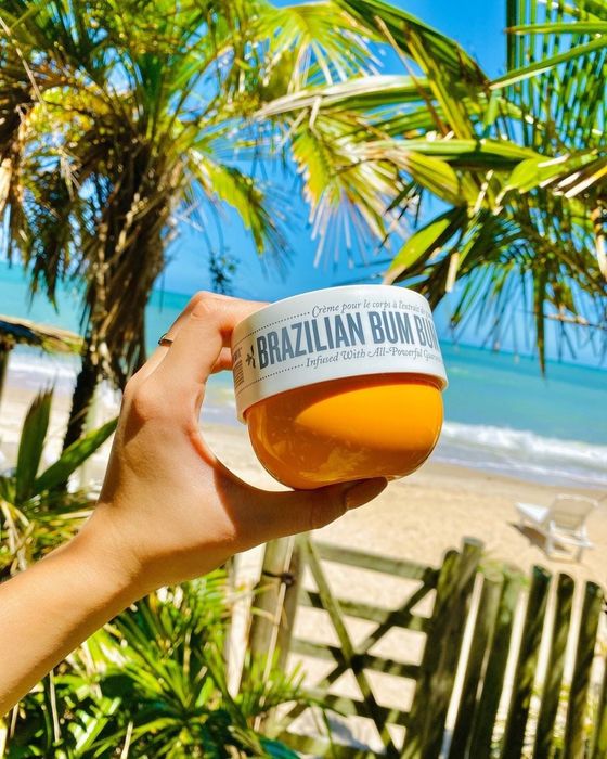 Model holding a jar of Brazilian Bum Bum Cream while lounging on a tropical beach