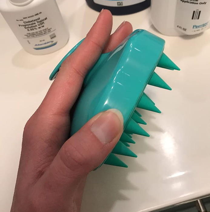 A hand holding a teal scalp massager, surrounded by various anti-dandruff shampoos and other hair care products.